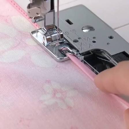 A close-up of a sewing machine presser foot guiding light pink fabric with a white floral pattern, with a finger holding a matching pink ribbon or guide in place.