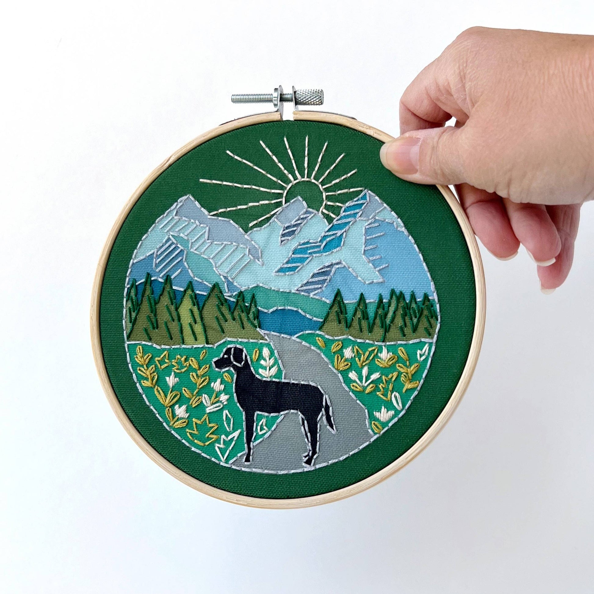 Embroidery hoop with a scenic design of mountains, trees, and a dog, held by a hand against a white background.