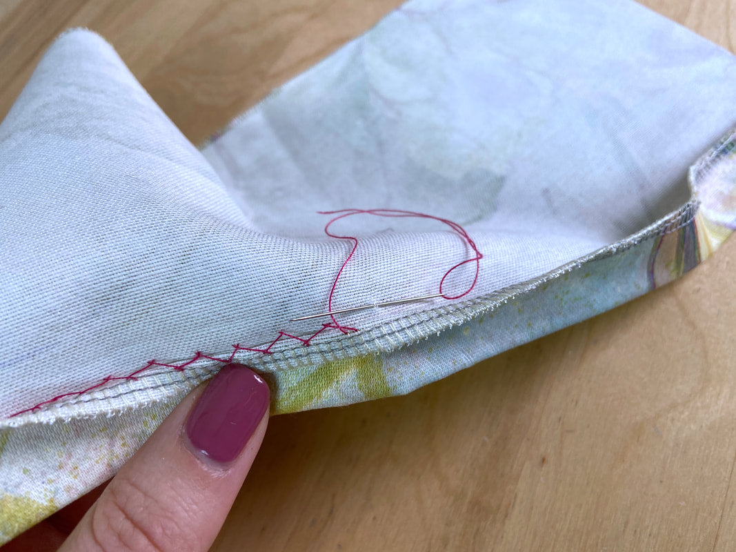 A close-up of a hand with magenta nail polish holding a folded piece of white fabric with a subtle pastel print and red stitching, set against a light wood background.
